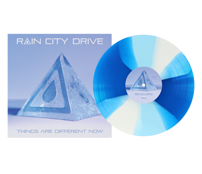 A transparent royal blue and white limited edition vinyl by Rain City Drive sits beside its full-color sleeve, which shows a glass pyramid holding a water droplet. The cover reads "RAIN CITY DRIVE" and "THINGS ARE DIFFERENT NOW.